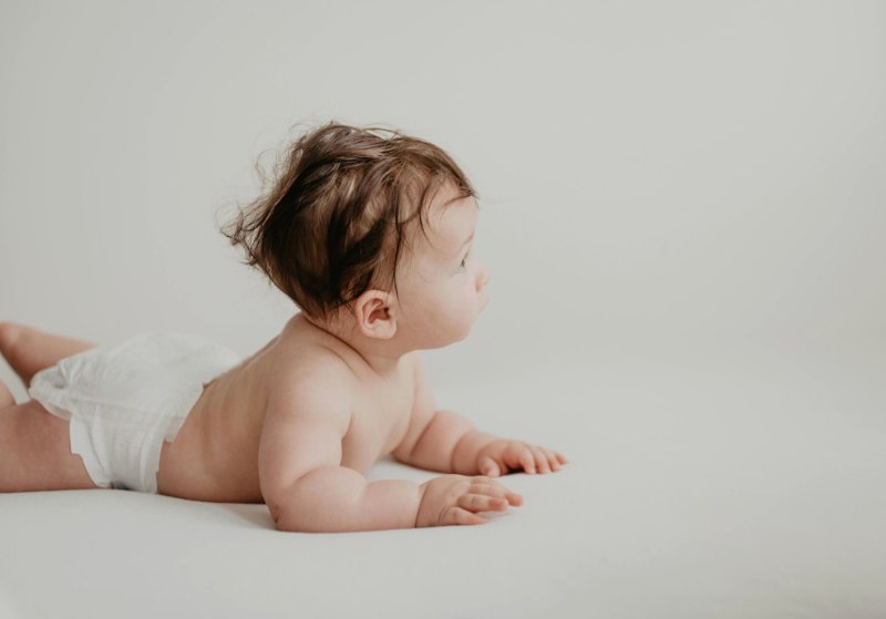 A baby lying on their front on a pale sheet, lifting their head and looking to the side
